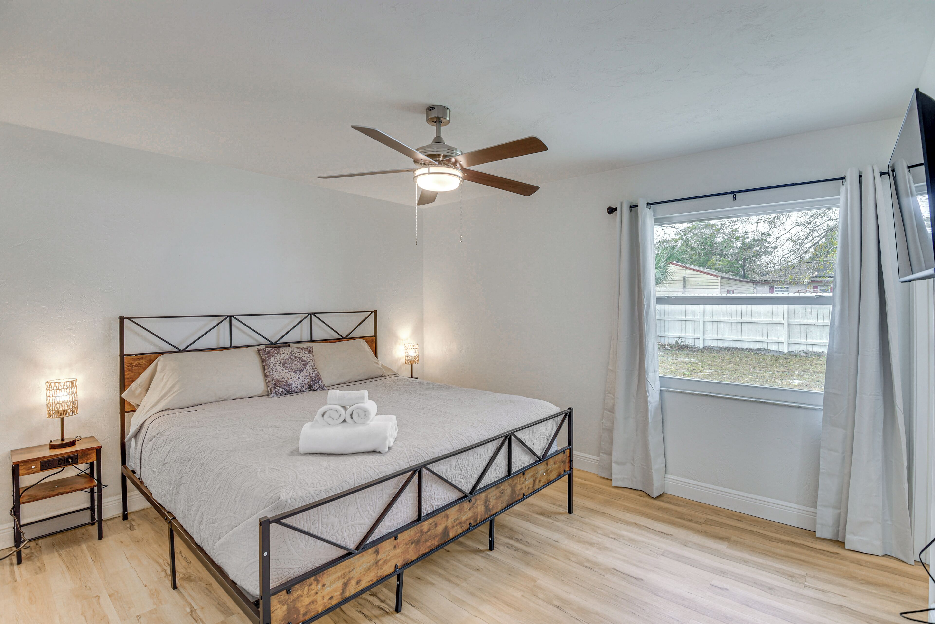A bedroom with a bed, a ceiling fan, a nightstand, a window with curtains, and a view of the outside.