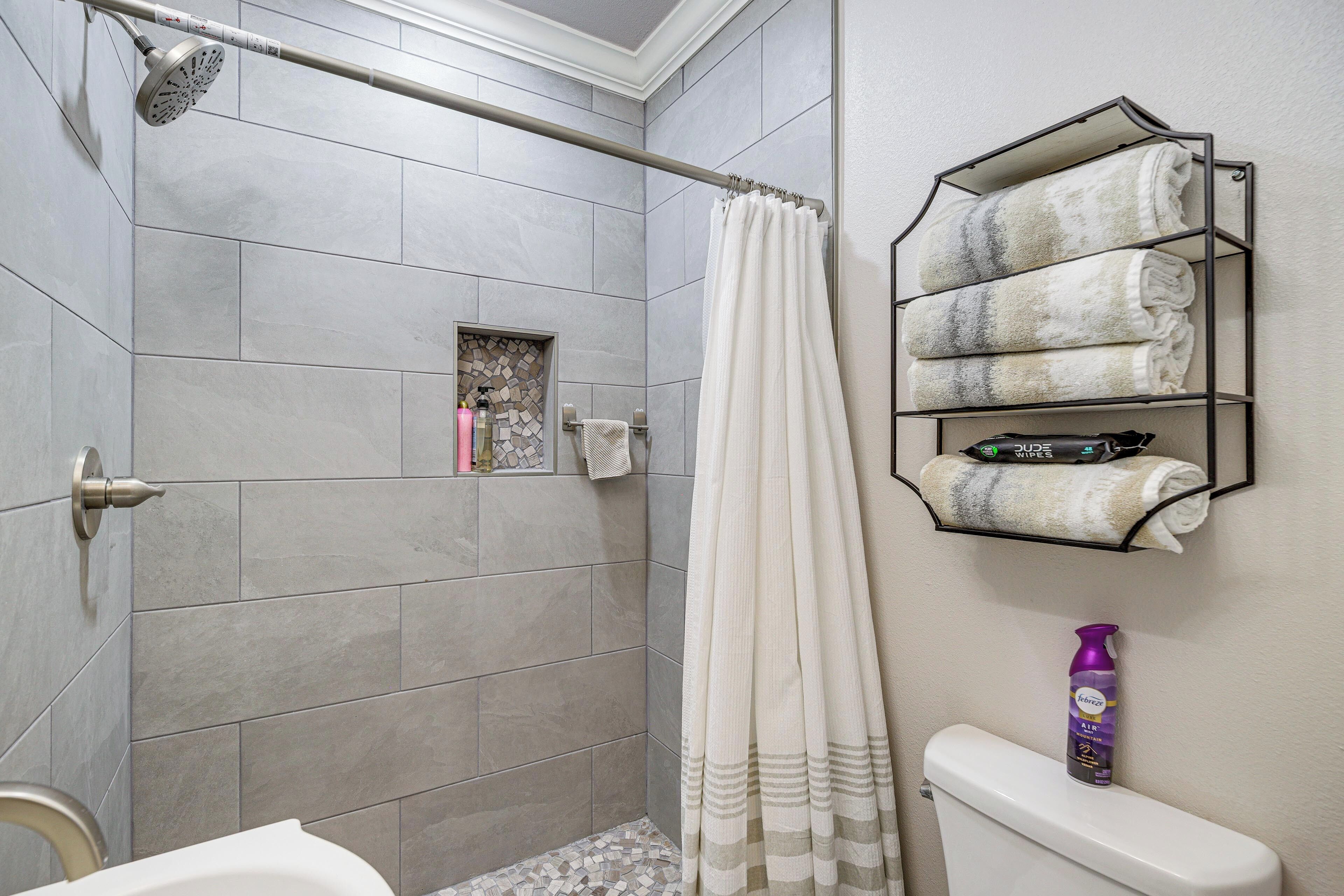 Un baño con azulejos grises, una ducha con cortina, un inodoro y un estante con toallas.