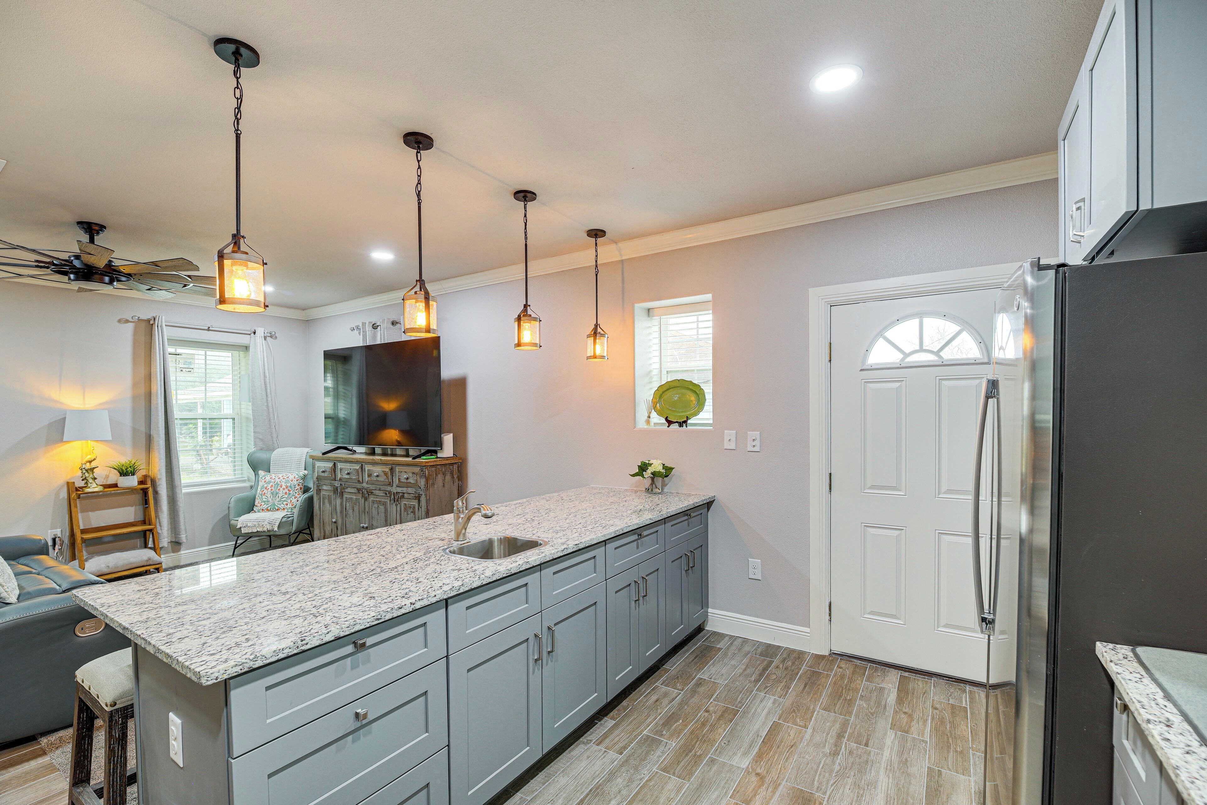 A kitchen with granite countertops, grey cabinets, a stainless steel refrigerator, pendant lights, and a ceiling fan.