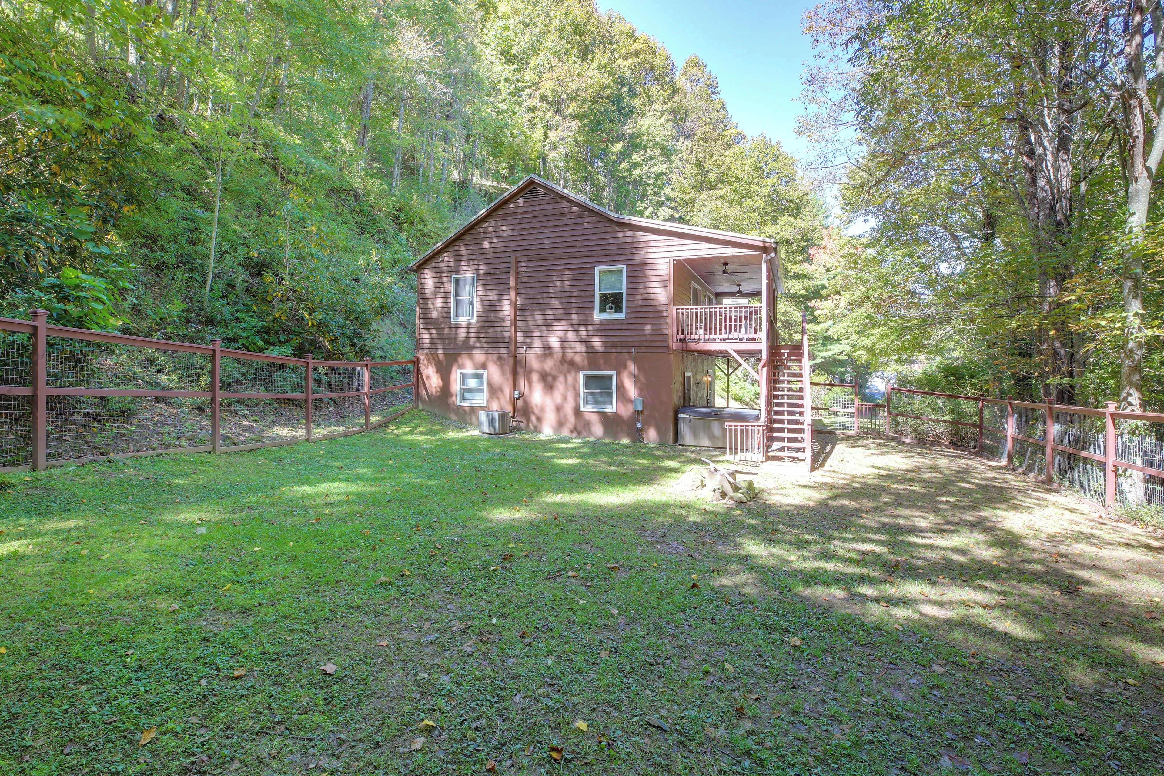 A two-story wooden cabin with a balcony, surrounded by a fenced yard and dense forest.