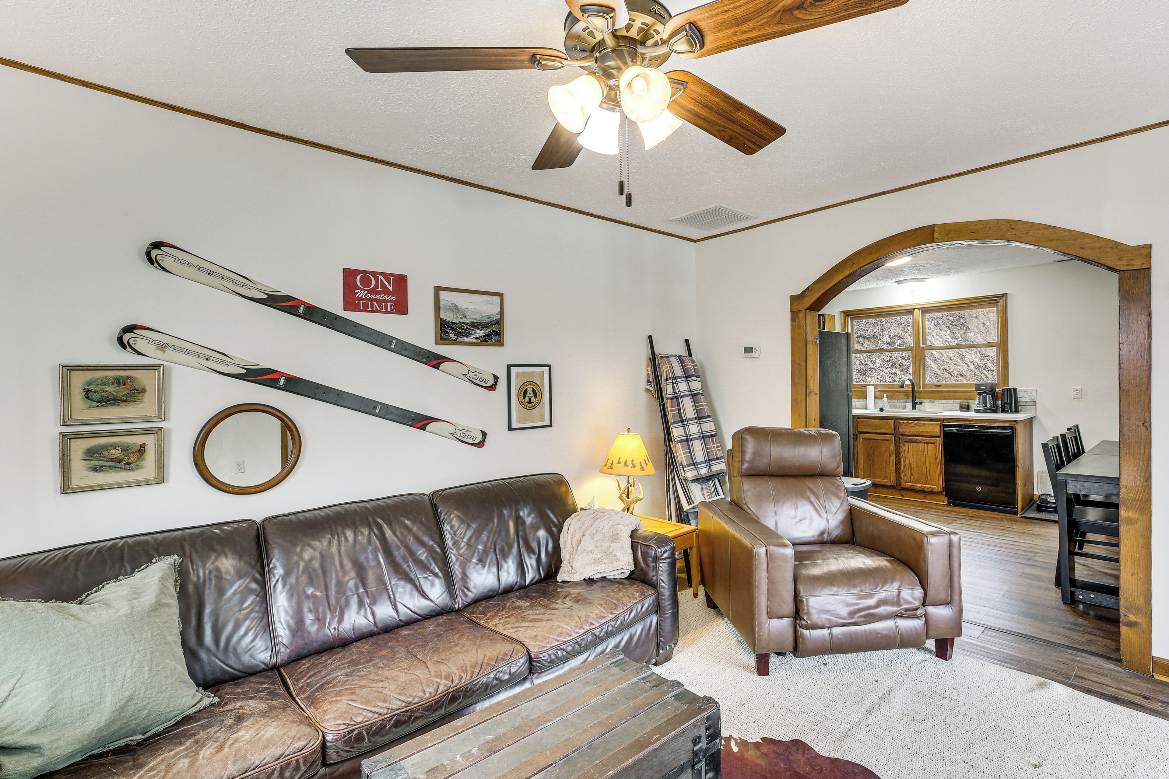A living room with a brown leather sofa, a brown leather armchair, a ceiling fan, and skis mounted on the wall.