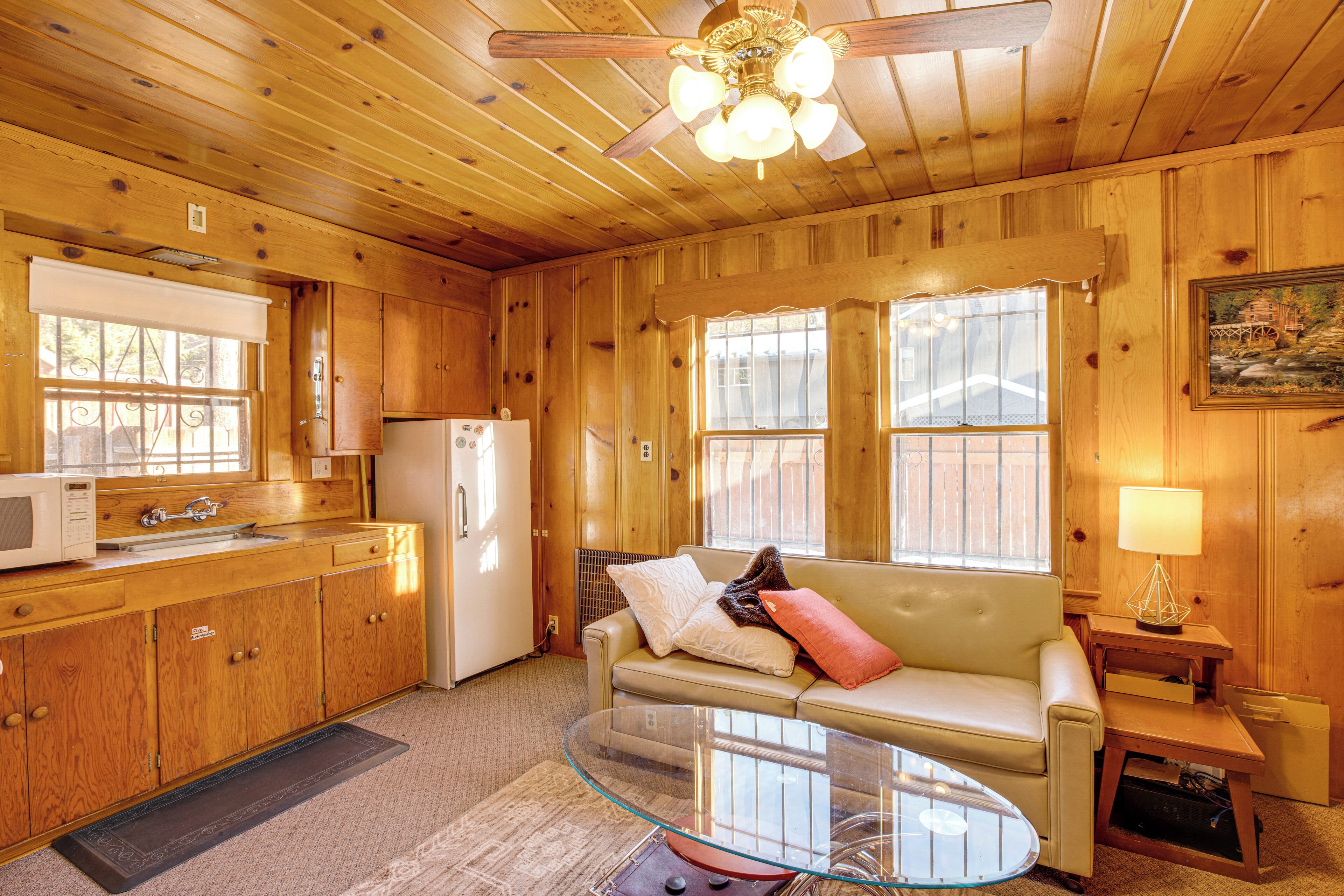 A wooden cabin interior with a kitchenette, a glass coffee table, a sofa with cushions, and a ceiling fan.
