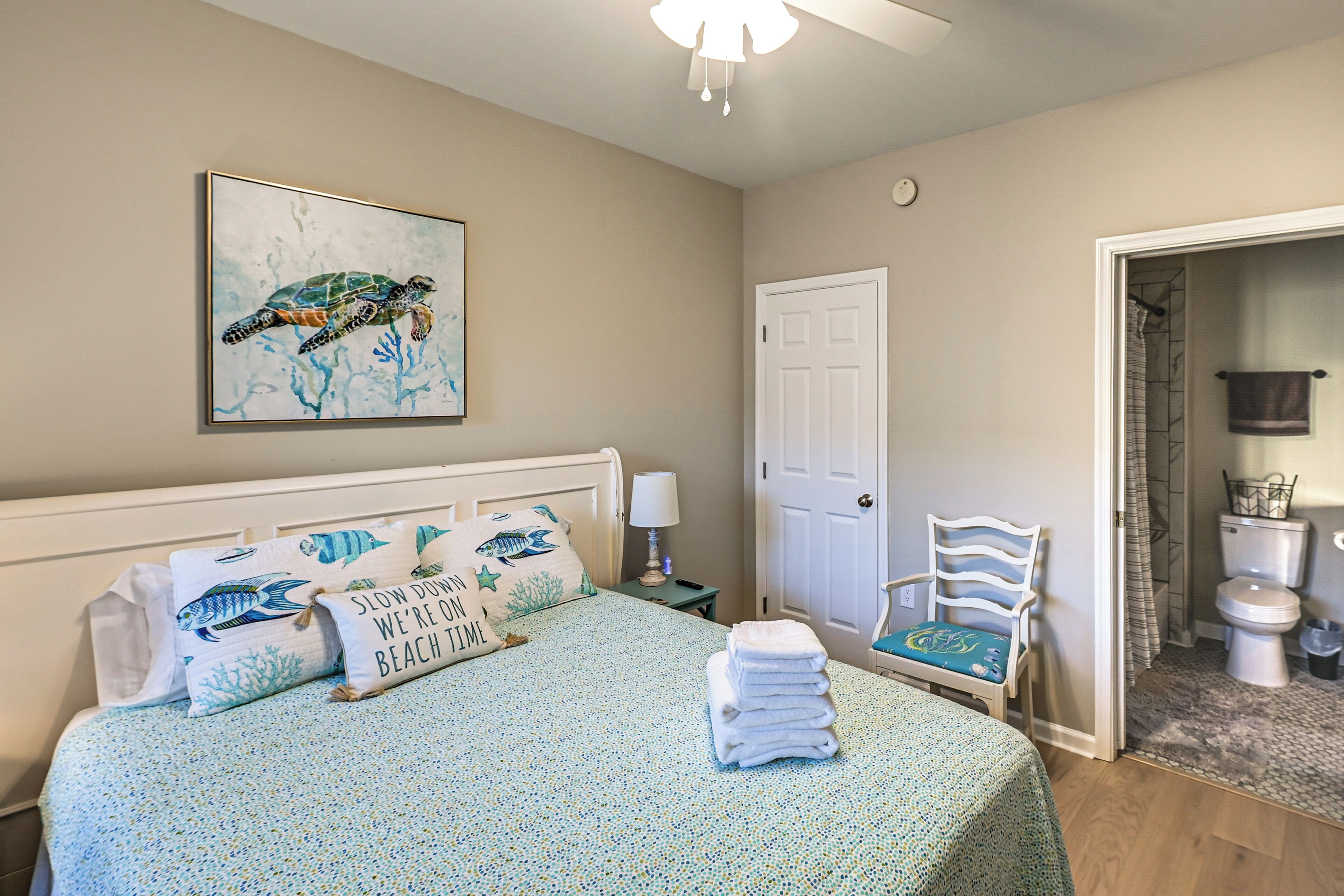 A bedroom with a bed, a nightstand, a chair, a door, and a bathroom visible through an open door.
