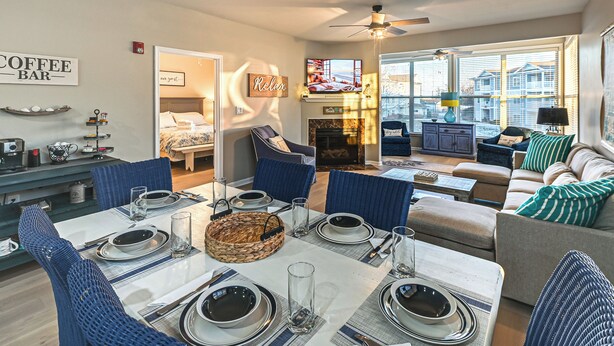 A modern living room with a dining area set for a meal, a fireplace, and a large window offering a view of the city. - Bay-n-beach! Comfy Coastal Condo in Lewes! (Lewes)