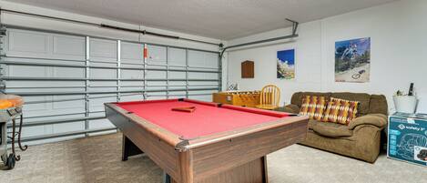 Une table de billard dans une pièce qui comprend une porte de garage, un canapé et une affiche accrochée au mur.