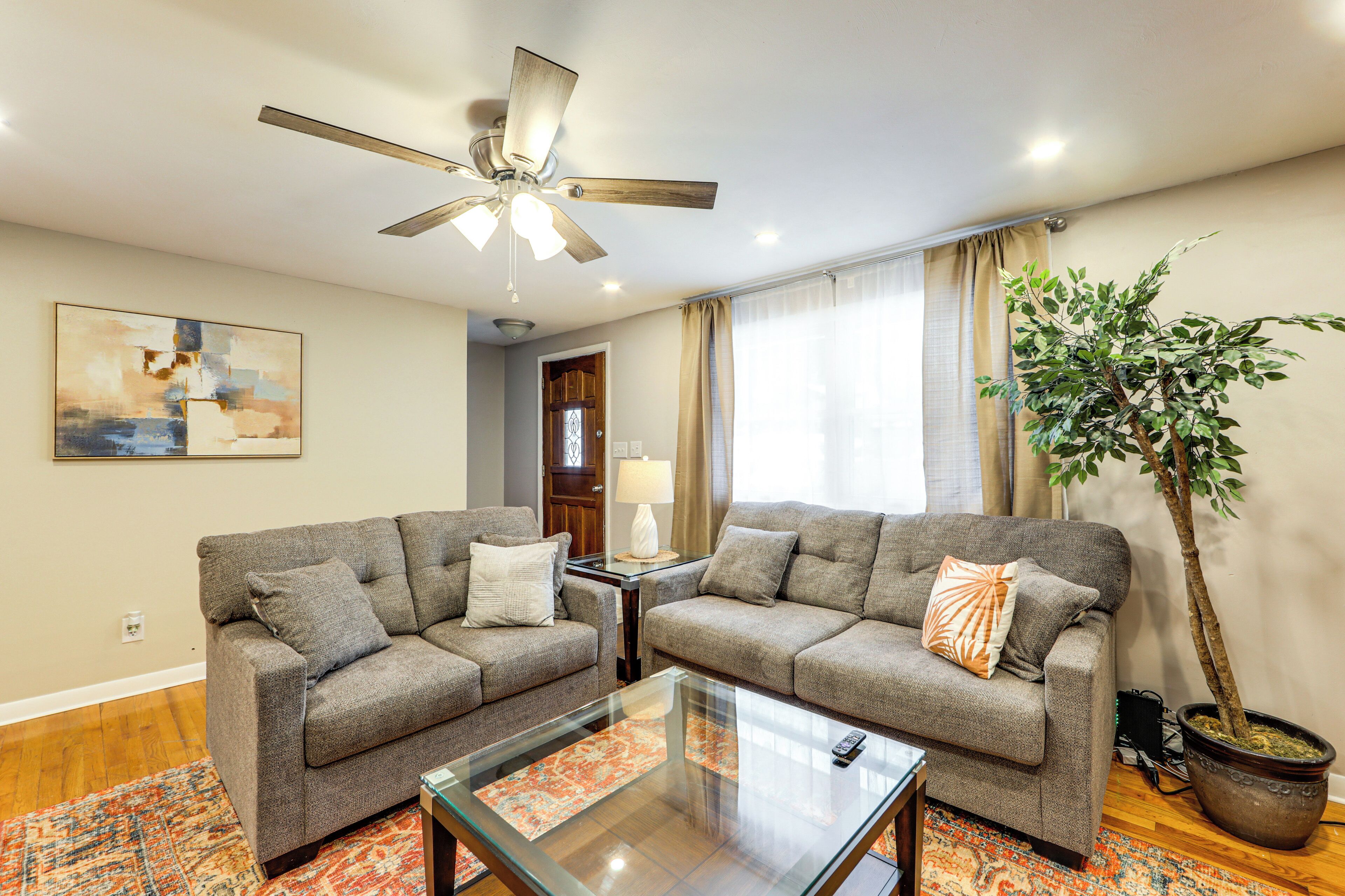 A living room with two grey sofas, a glass coffee table, a ceiling fan, a potted plant, and a large abstract painting on the wall.