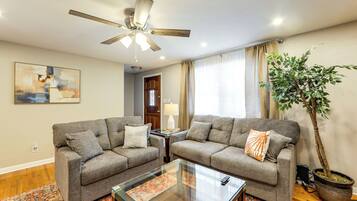 A living room with two grey sofas, a glass coffee table, a ceiling fan, a potted plant, and a large abstract painting on the wall.