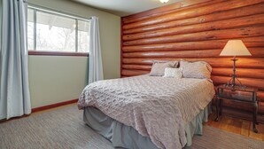 Un dormitorio con pared de troncos de madera, cama con manta estampada, mesita de noche con lámpara y ventana con cortinas.