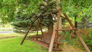 Un ensemble de balançoires en bois rustique, installé dans un jardin avec un grand arbre et un mur de pierre en arrière-plan.
