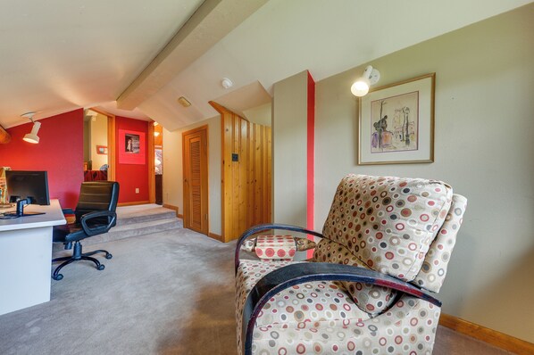 Una habitación con un escritorio, una silla de oficina y un sillón con estampado. Hay un cuadro en la pared y una puerta que da a otra habitación.