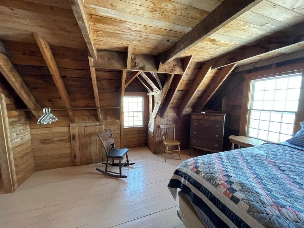 A rustic attic bedroom with exposed wooden beams, a rocking chair, a dresser, and a quilted bedspread.