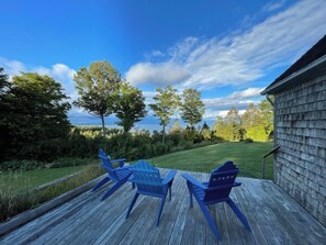 A wooden deck with two Adirondack chairs and a view of a grassy area and trees. - Private Beach! Passamaquoddy Bay Cottage (Perry)