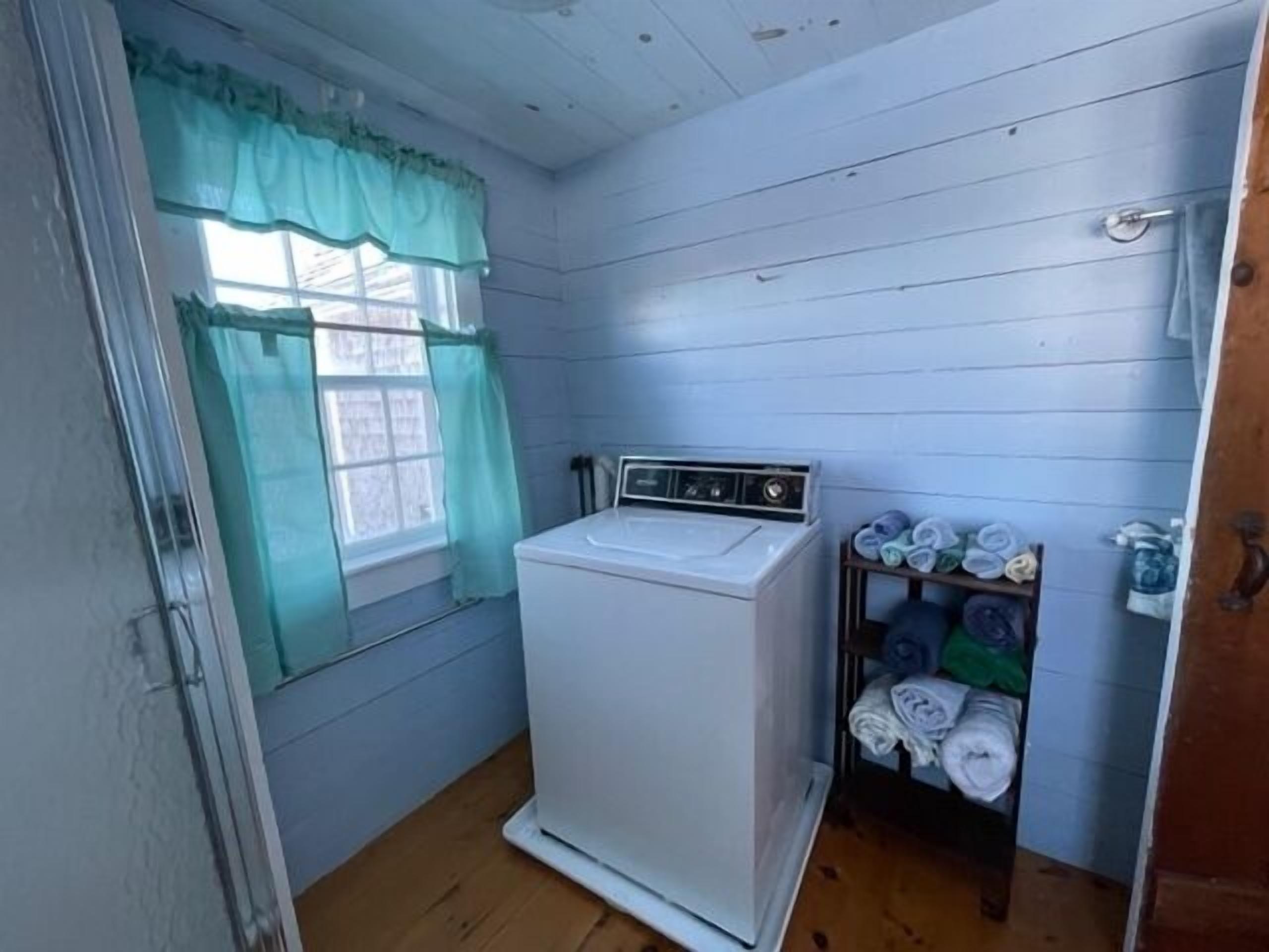 A laundry room with a washing machine, a window with a green curtain, and a shelf with folded towels.