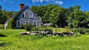A two-story house with a gabled roof and a chimney, surrounded by a well-maintained lawn and a stone wall. - Private Beach! Passamaquoddy Bay Cottage (Perry)