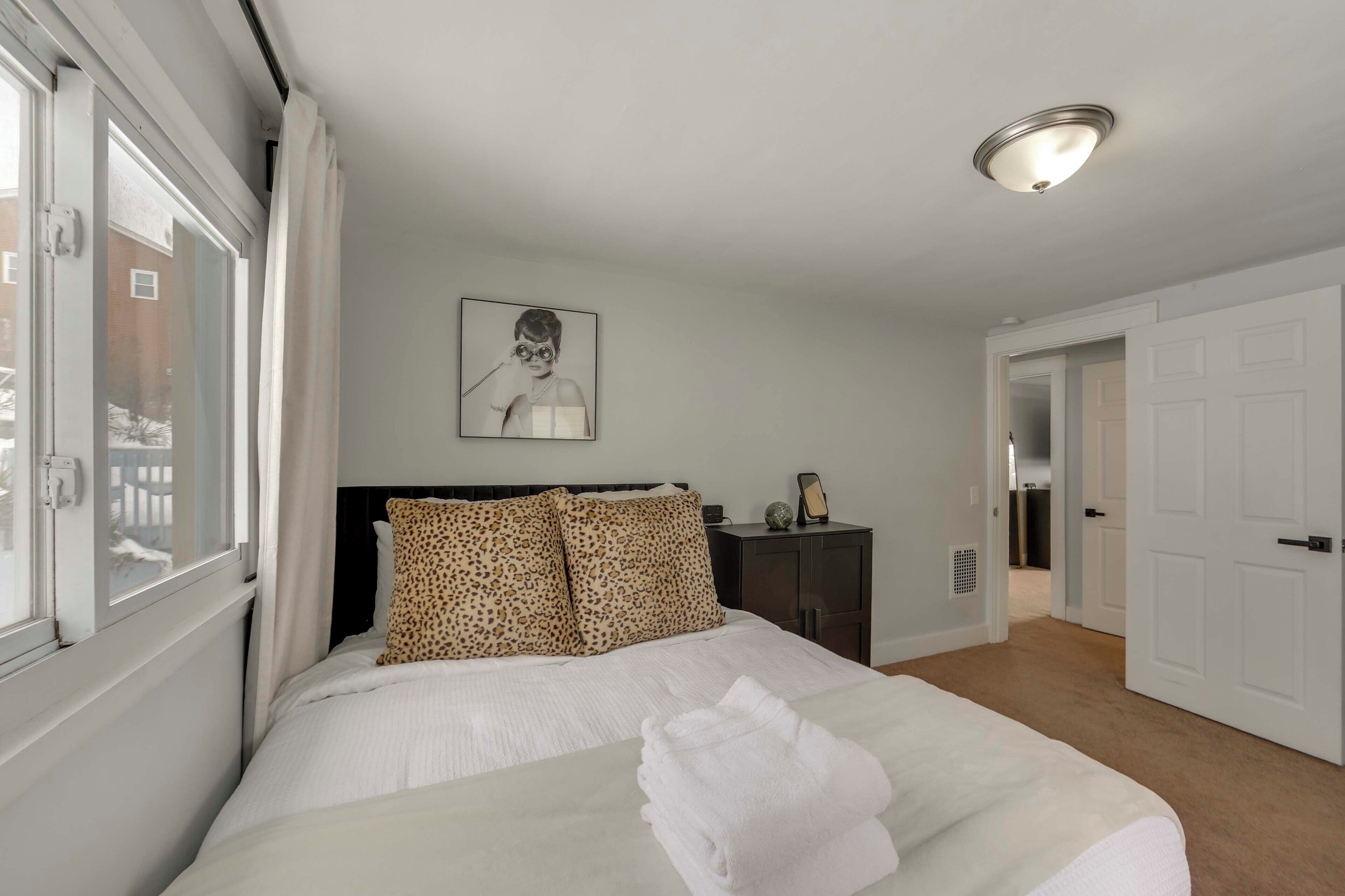 A neatly arranged bedroom with a bed, a headboard, a bedside table, a framed picture on the wall, and a door leading to another room.