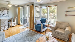 A spacious living room with a ceiling fan, a large window with curtains, a blue armchair, a beige armchair, and a coffee table with a glass of milk. - Golfer's Getaway! Modern Diamondhead Condo (Diamondhead)