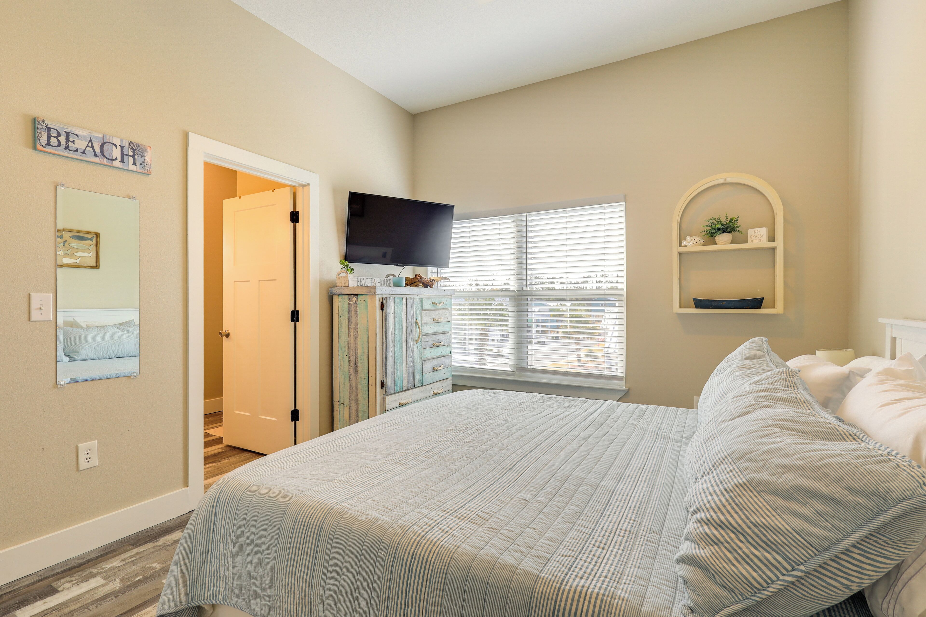 A bedroom with a bed, a television, a window with blinds, a dresser, and a wall-mounted shelf.