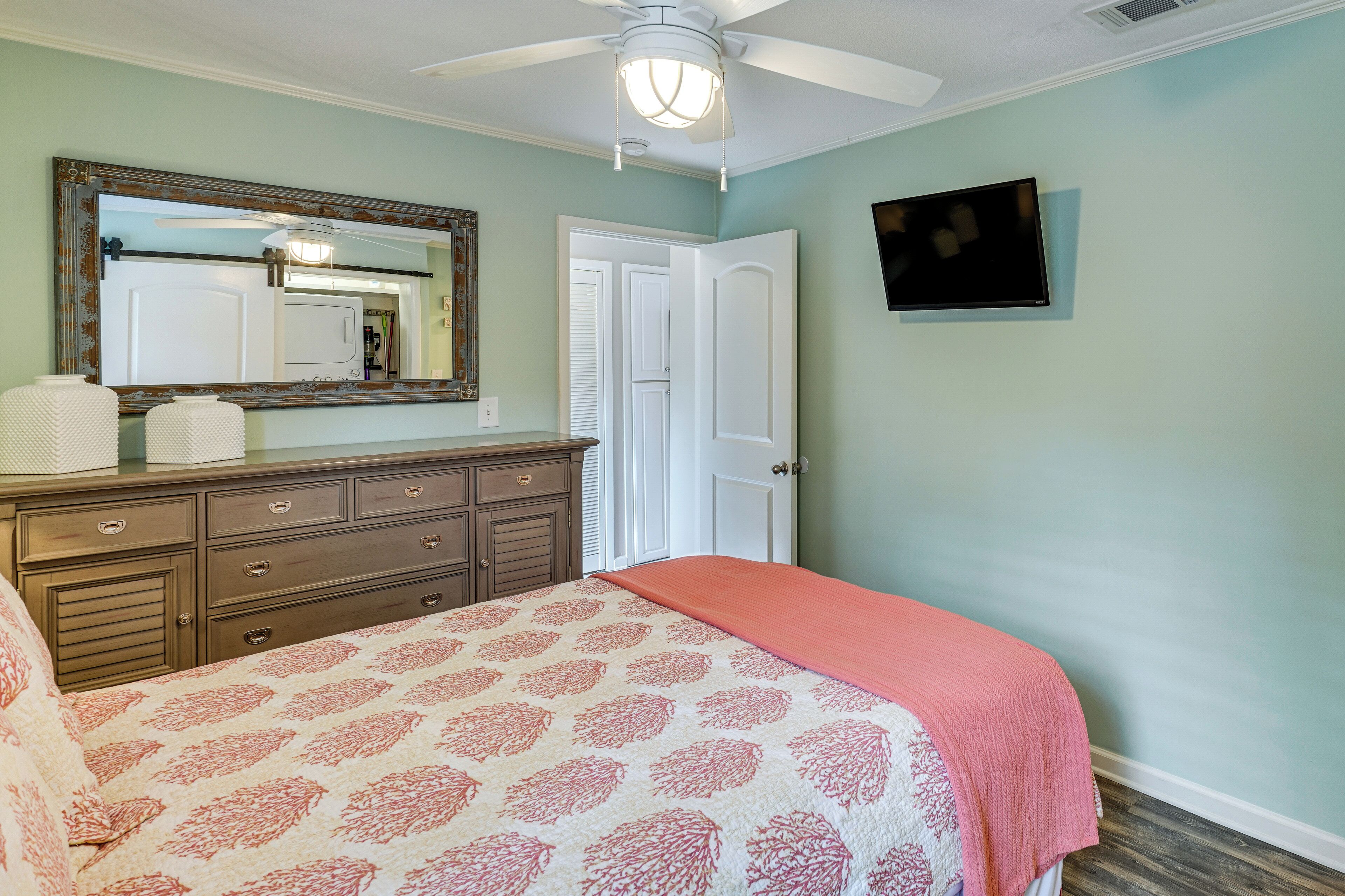 Une chambre à coucher comprenant un lit, une commode, un miroir, un ventilateur de plafond et un téléviseur fixé au mur.
