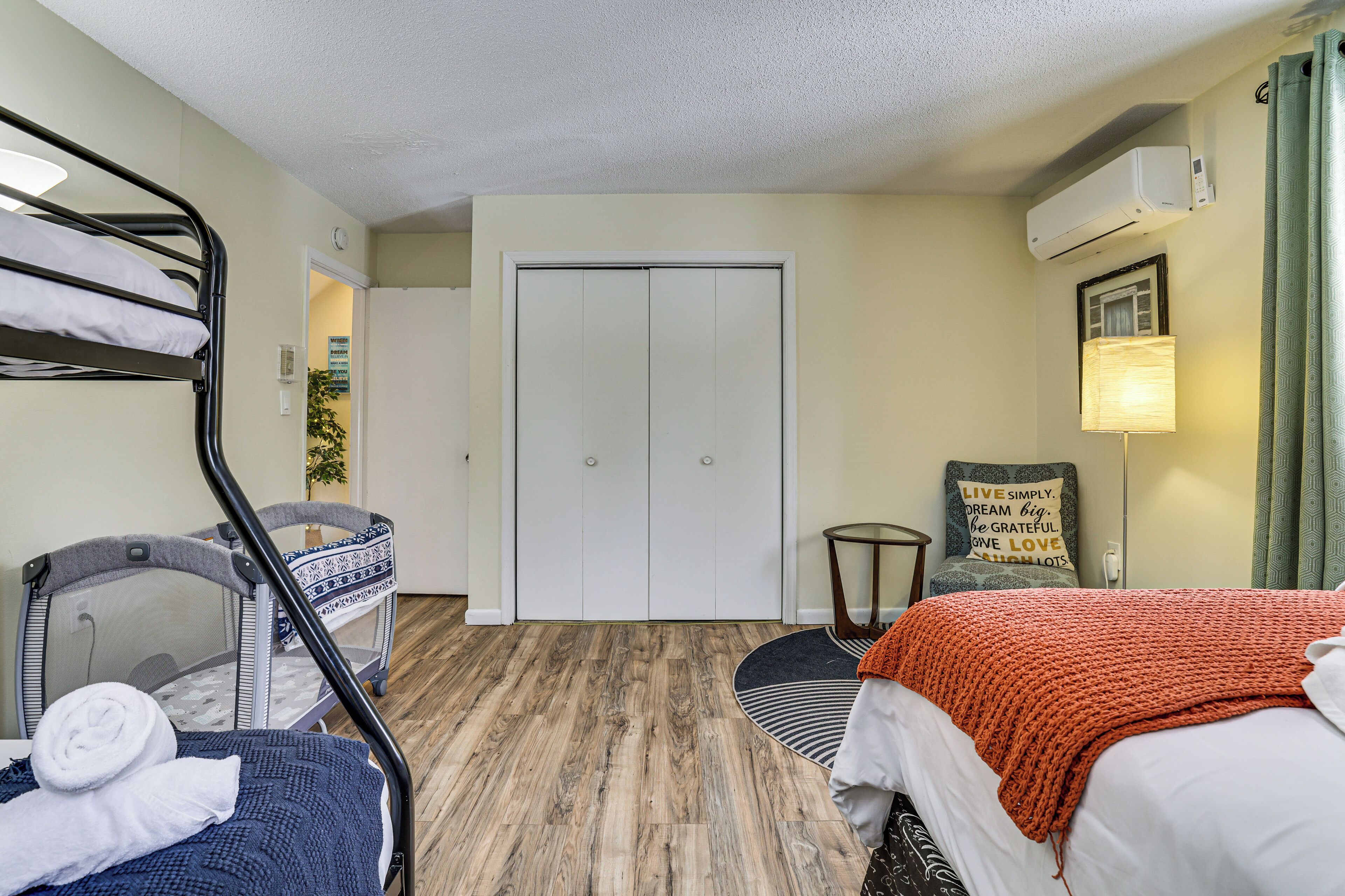 A hotel room with a bunk bed, a crib, a round rug, a small table, and a wall-mounted air conditioner.