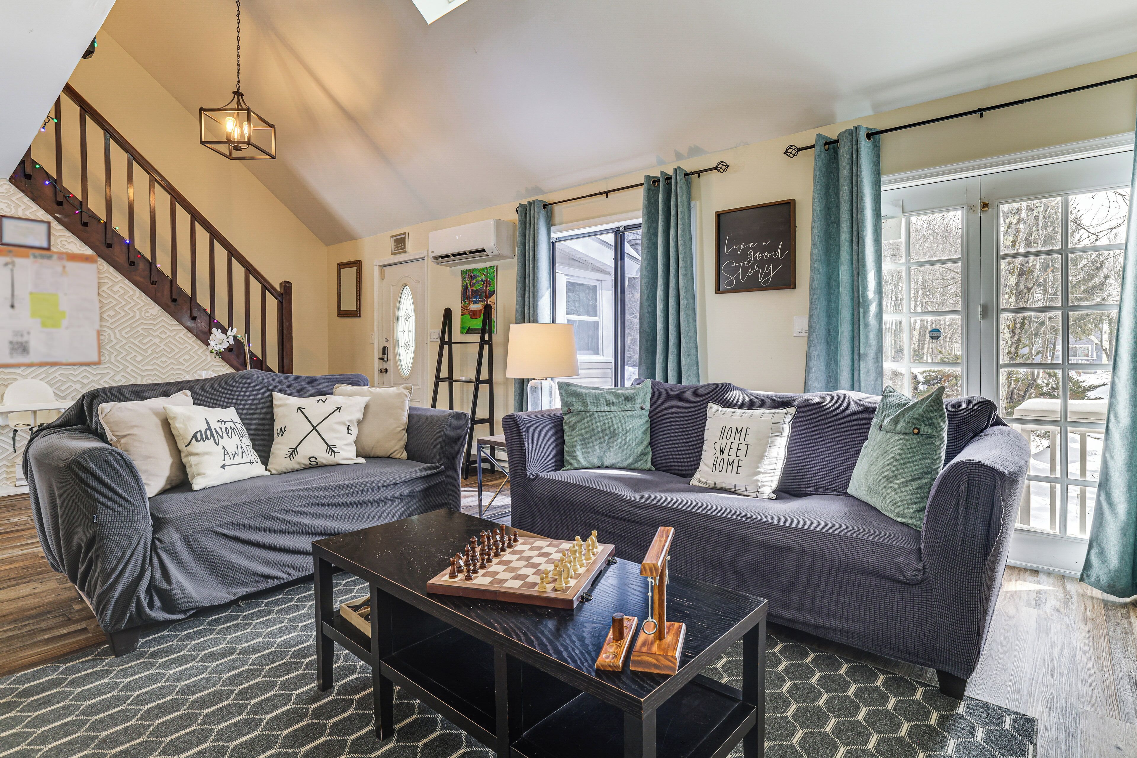 A living room with two sofas, a coffee table with a chess set, a staircase, and a large window with curtains.