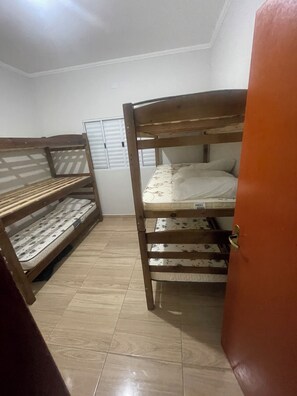 Um dormitório com beliches, piso ladrilhado e uma porta.