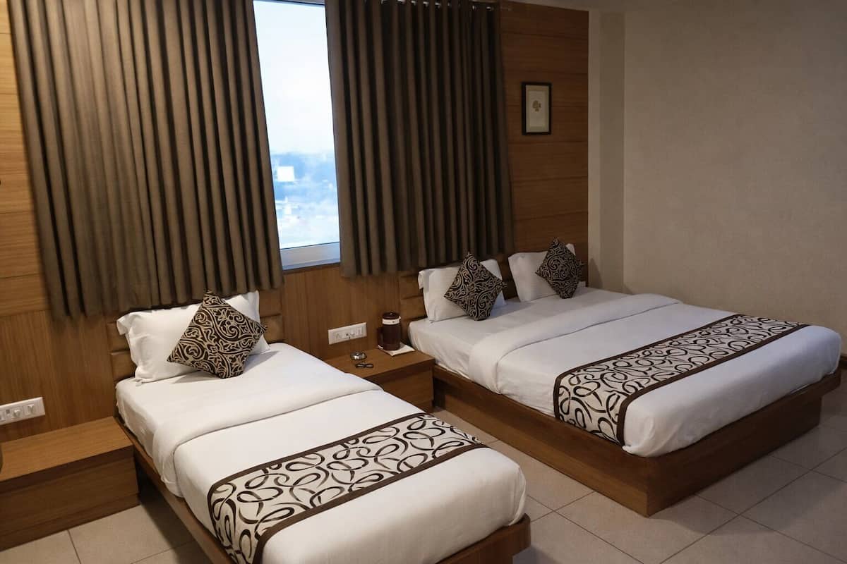 A hotel room with two beds, each with patterned bedspreads, a bedside table with a coffee maker, and a window with curtains.