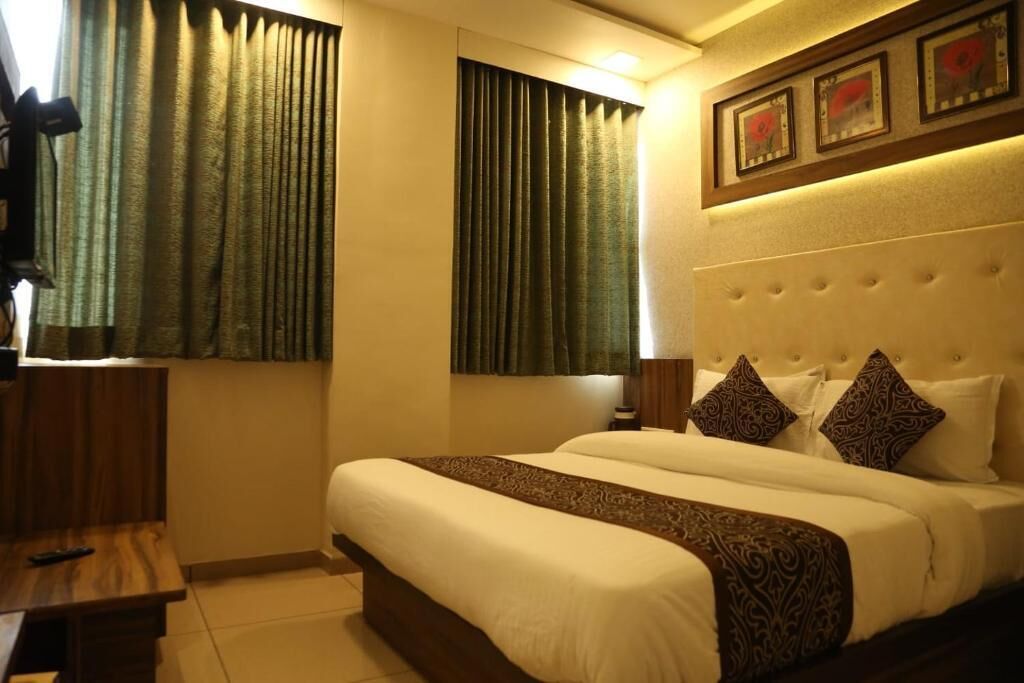 A hotel room with a large bed, two windows with curtains, a headboard with decorative pillows, and a framed picture above the bed.