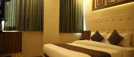 A hotel room with a large bed, two windows with curtains, a headboard with decorative pillows, and a framed picture above the bed.