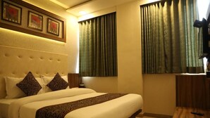 A hotel room with a large bed, two pillows, a headboard, a bedside table, a lamp, and a window with curtains. - Hotel RK Palace (Gandhinagar)