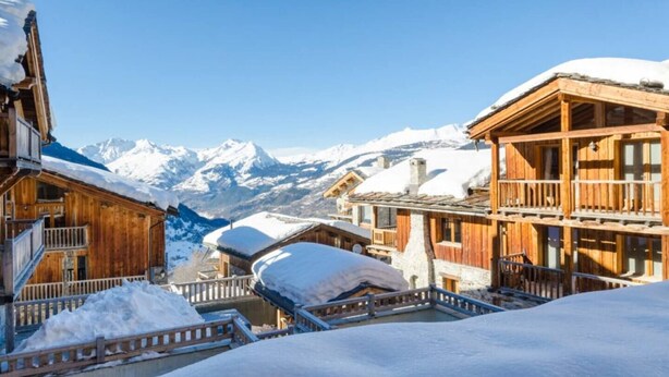 Een houten chalet met balkon, omringd door besneeuwde bergen en heldere luchten.