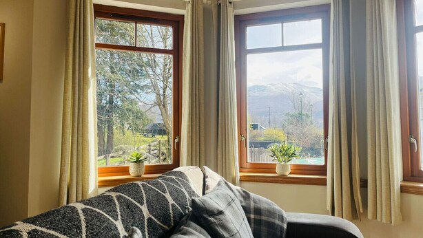 A room with a sofa, two windows with curtains, and a view of greenery outside.