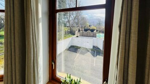 A window with a view of a paved area and a low wall, curtains partially drawn, and a plant at the window base.