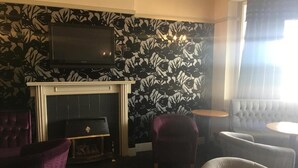 A room with a fireplace, a television mounted above it, and a patterned wall. There are two armchairs and a round table.