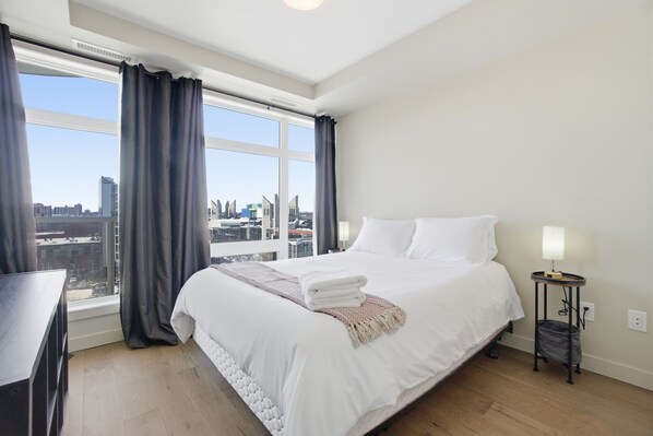 A hotel room with a large window, a bed with white linens, a bedside table with a lamp, and a small trash bin.