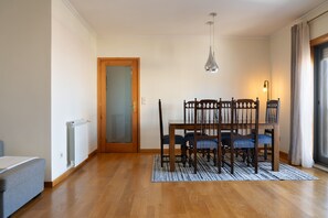 Un comedor con mesa y sillas de madera, un radiador, un sofá gris y una puerta que da a otra habitación.