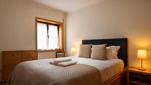 A neatly made bed with a quilted cover, two bedside lamps, a window with curtains, and a wooden nightstand.