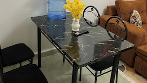 Un'area pranzo con un tavolo in marmo nero, un distributore di acqua, un vaso con fiori gialli, un telecomando e un quadro appeso al muro.
