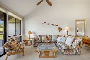 A living room with a sectional sofa, a glass coffee table, and a ceiling fan. - Peaceful Golf Course Retreat with Partial Ocean Views-Wailea Grand Champions #78 (Wailea, Maui)