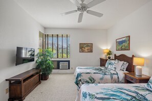 A bedroom with a bed, a TV, a ceiling fan, a window with blinds, a plant, and a painting on the wall. - Peaceful Golf Course Retreat with Partial Ocean Views-Wailea Grand Champions #78 (Wailea, Maui)