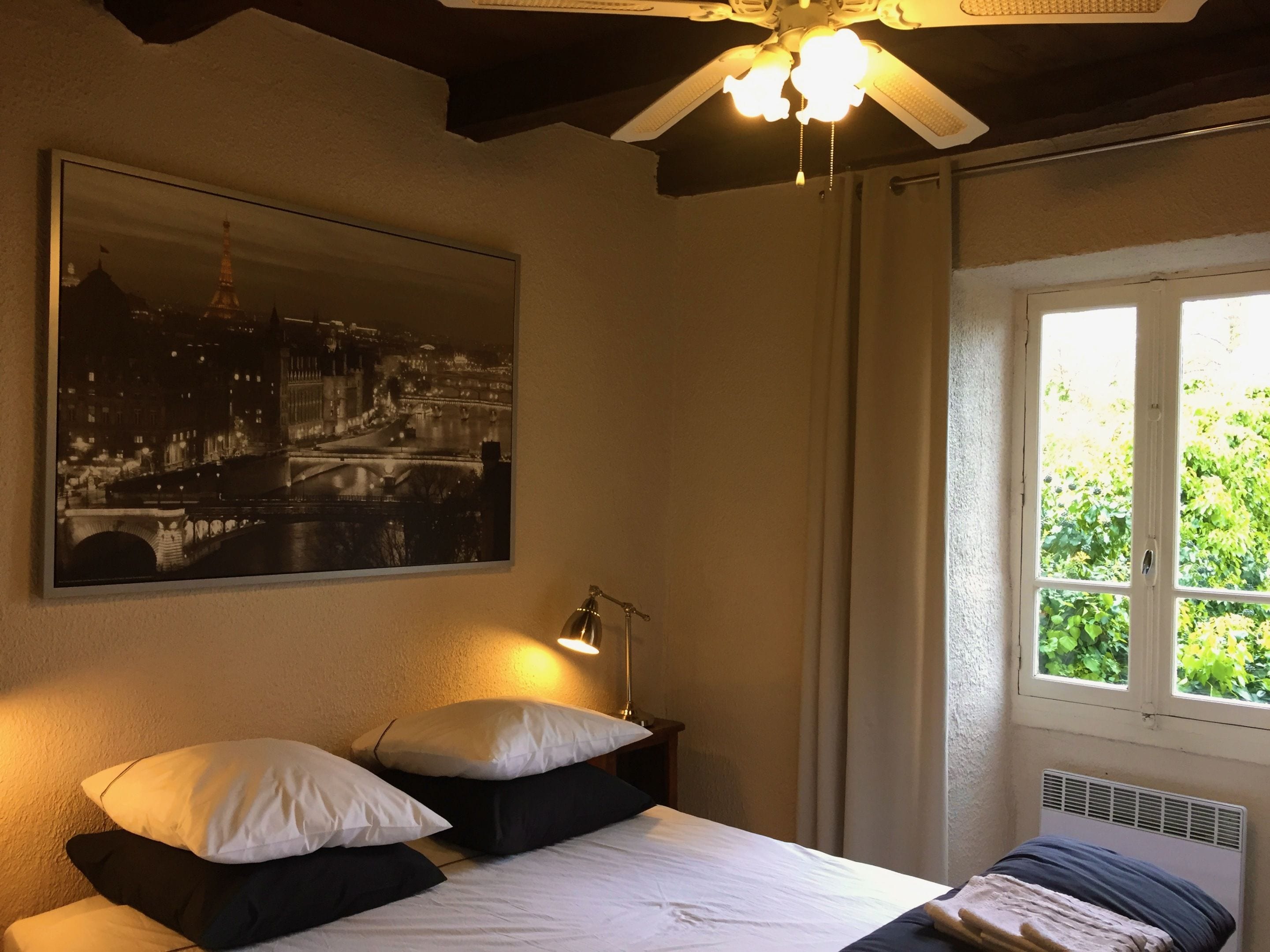 A bedroom with a bed, pillows, a nightstand, a lamp, a ceiling fan, a window with curtains, and a cityscape painting on the wall.