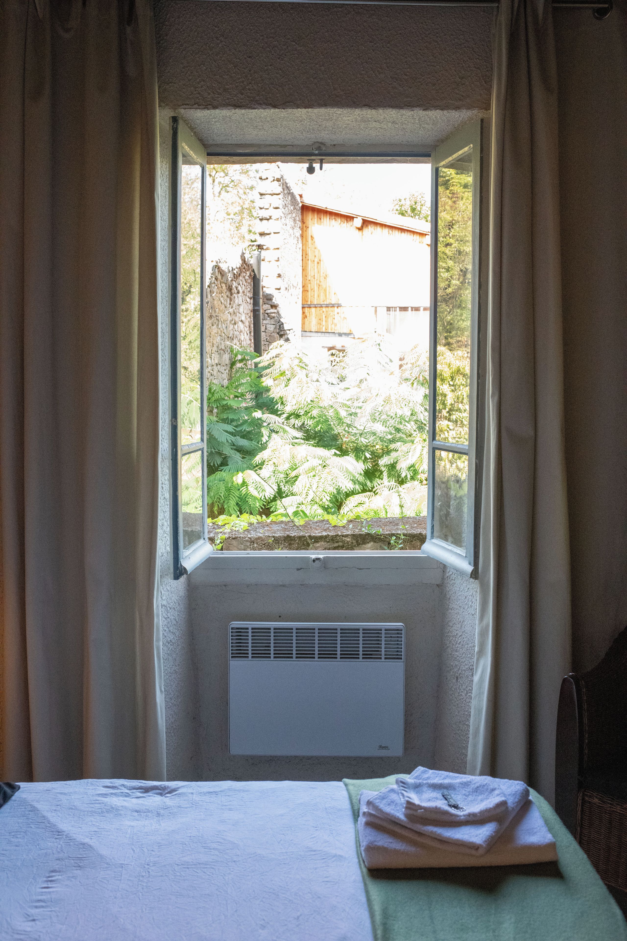 A room with a window overlooking a garden, a radiator, and a bed with a folded towel.