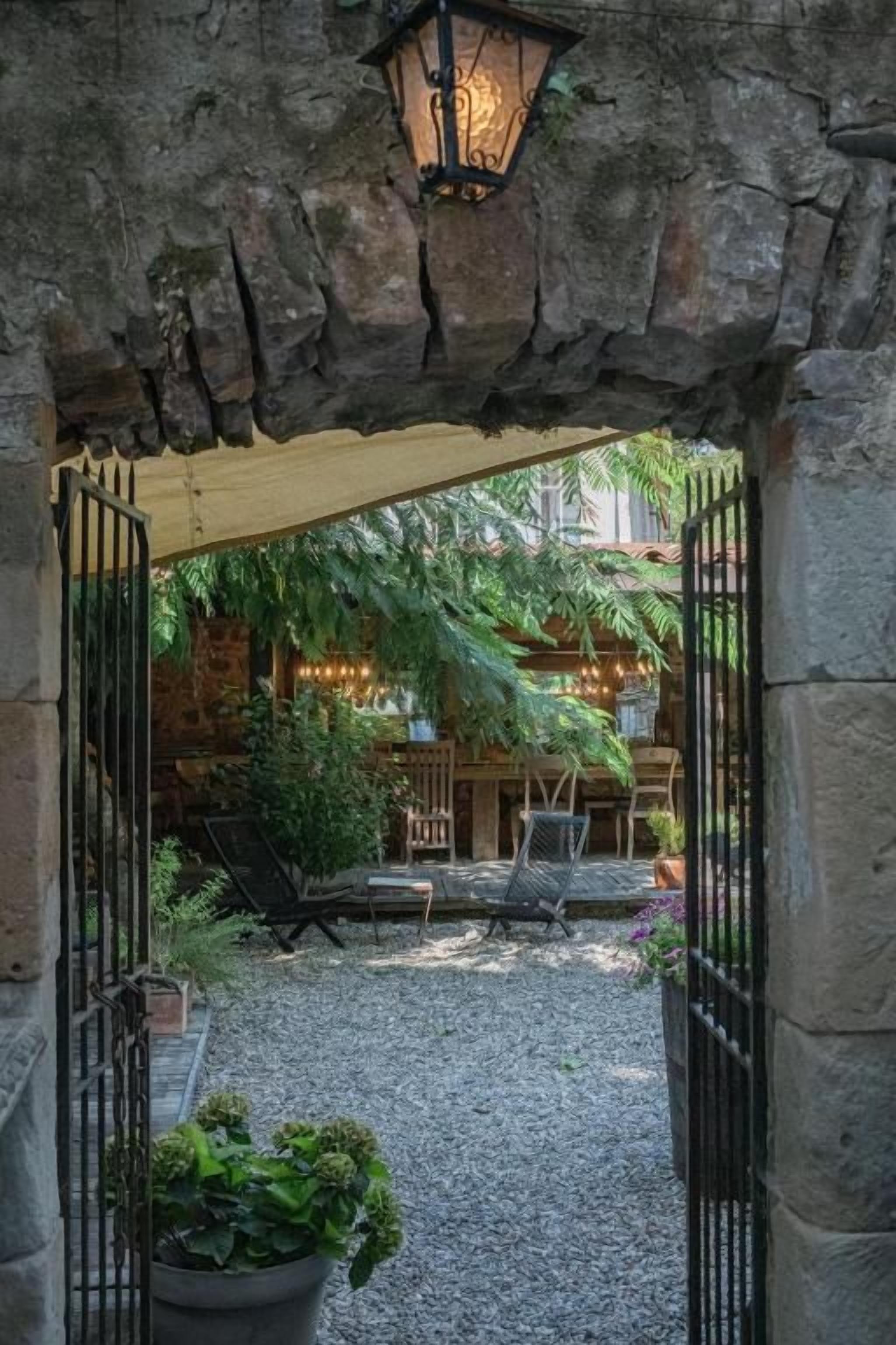 An outdoor patio area with a stone archway, a lantern, and a seating area with a view of a building with a wooden facade.