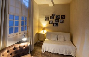 A small, cozy bedroom with a single bed, a bedside table with a lamp, a chair, and a wall with framed pictures.
