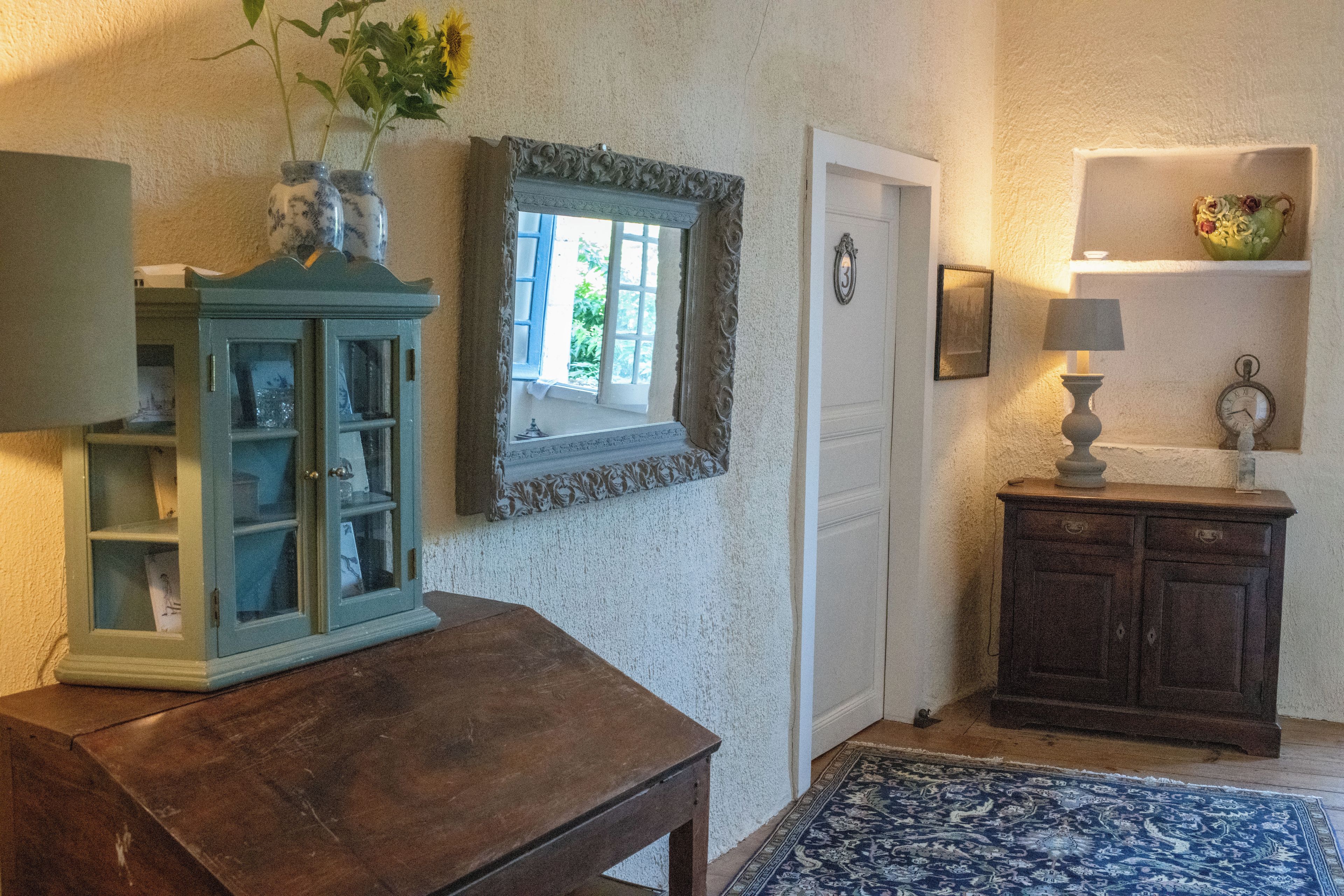 Une pièce comprenant une table en bois, un meuble avec des portes en verre, un miroir doté d’un cadre décoratif, une porte avec une plaque numérotée et une petite étagère sur laquelle se trouvent une lampe et une horloge.