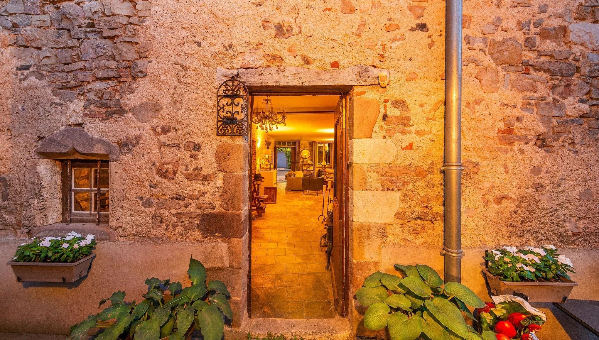 A stone building with a doorway leading to a well-lit interior. There are potted plants on either side of the entrance.