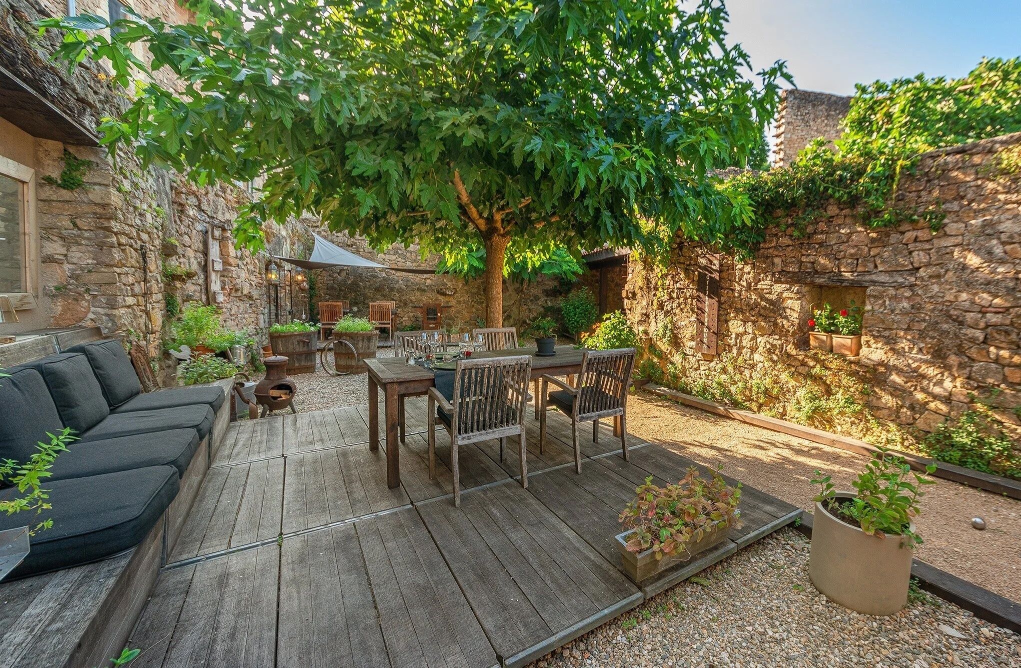 Un patio rústico al aire libre con mobiliario de madera, un muro de piedra y un árbol que da sombra.