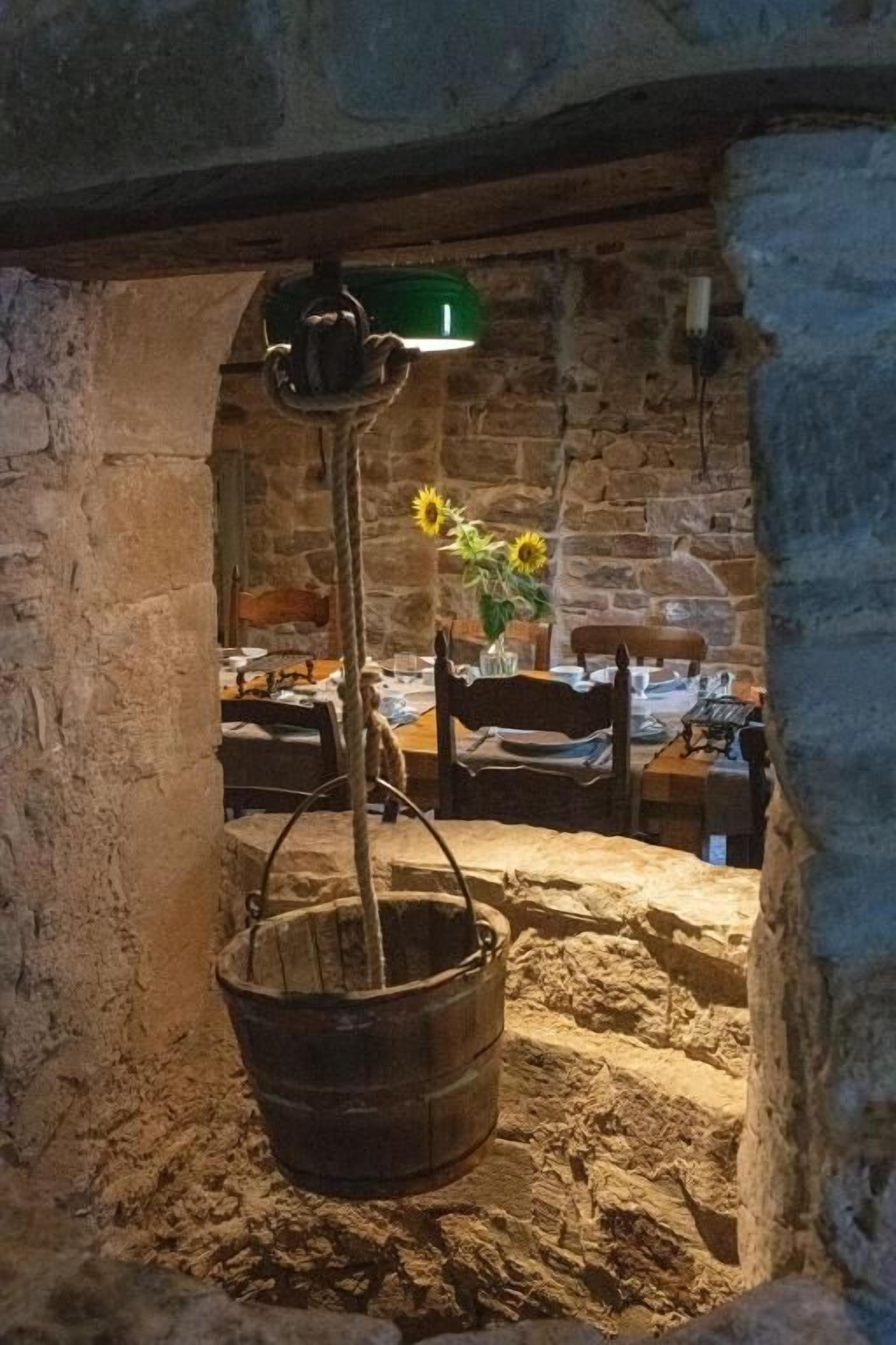 Una habitación rústica con pared de piedra, un balde de madera y una lámpara colgante.