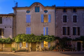 A historic building with a central entrance, a clock, and a garden area with potted plants. - La Maison au Puits (Cordes-sur-Ciel)