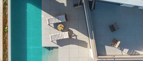 An aerial view of a swimming pool with two sun loungers and a small table with yellow balls, surrounded by a tiled deck.