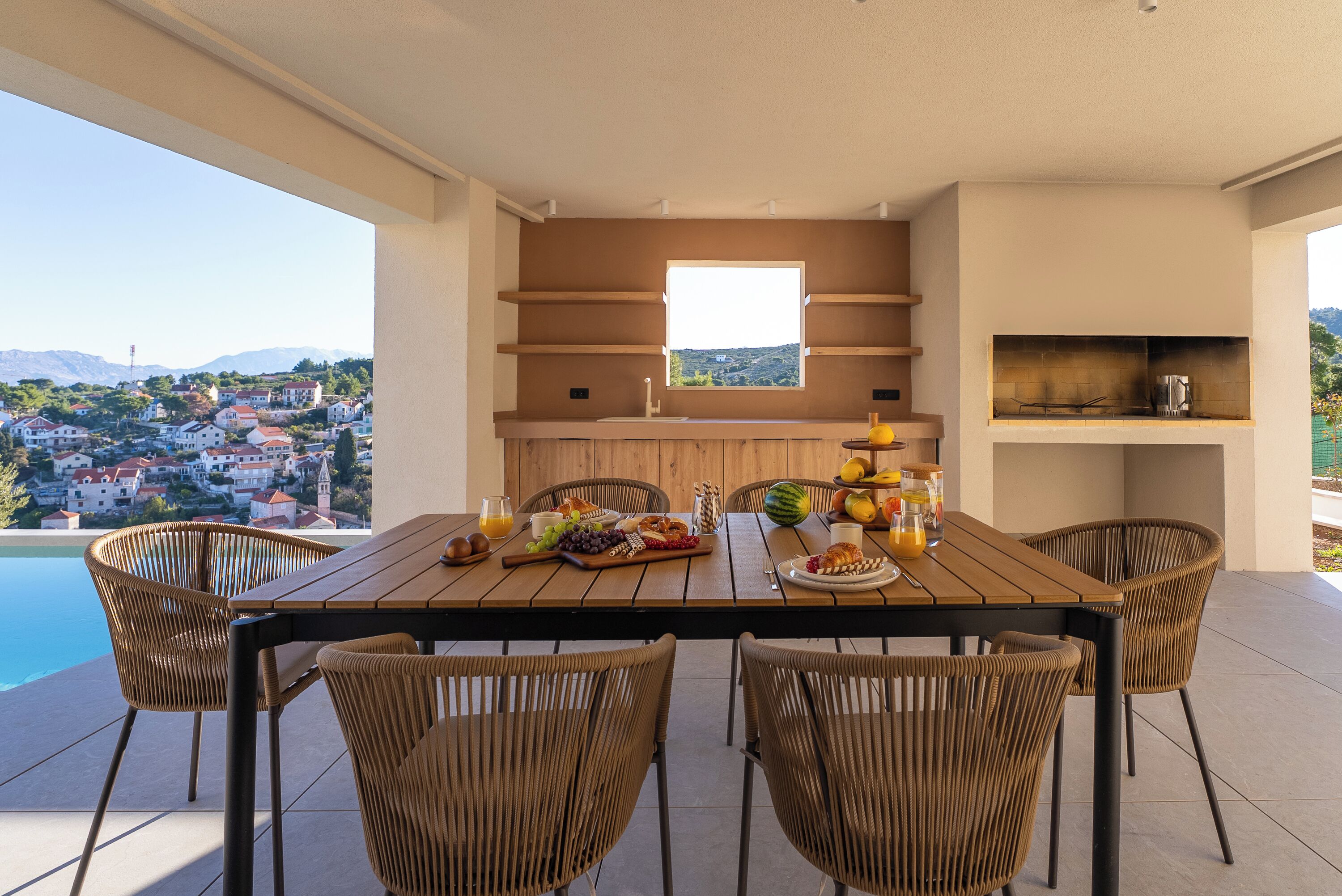 Un comedor con una mesa dispuesta para cuatro personas, rodeada de sillas de mimbre, con vista a una piscina y una panorámica de un pueblo.